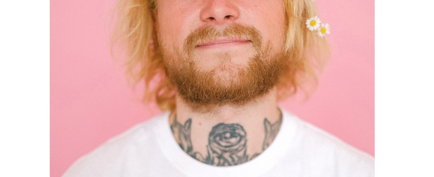Man with blonde hair and a beard, with a flower in his hair and a rose tattoo on his neck against a pink background.